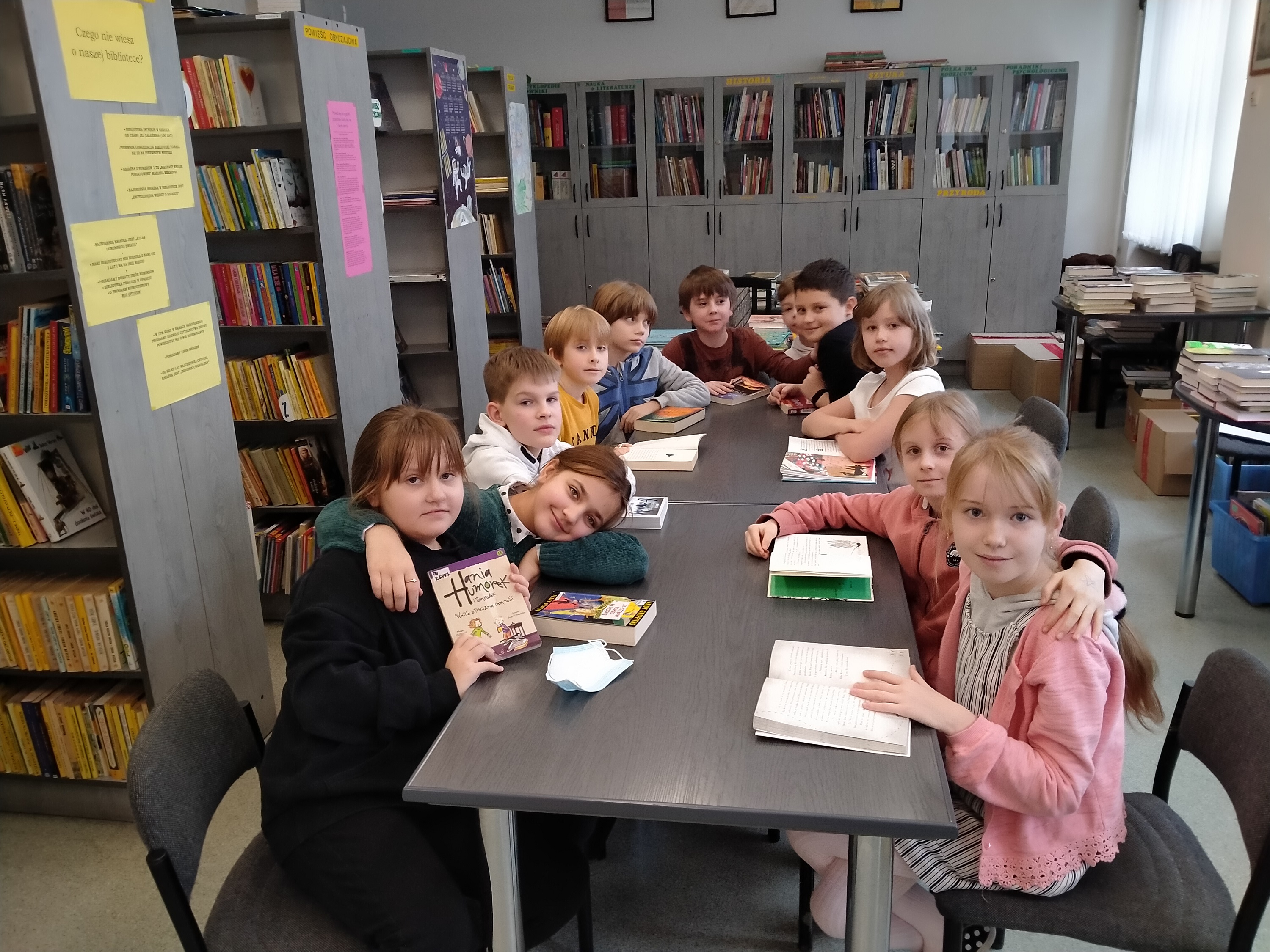 Grupa dzieci: pięć dziewczynek i 6 chłopców siedzi wokół dużego stołu w bibliotece szkolnej. Przed każdym z uczniów na stole leży otwarta książka. Dzieci patrzą w obiektyw. W tle znajdują się regały z książkami.