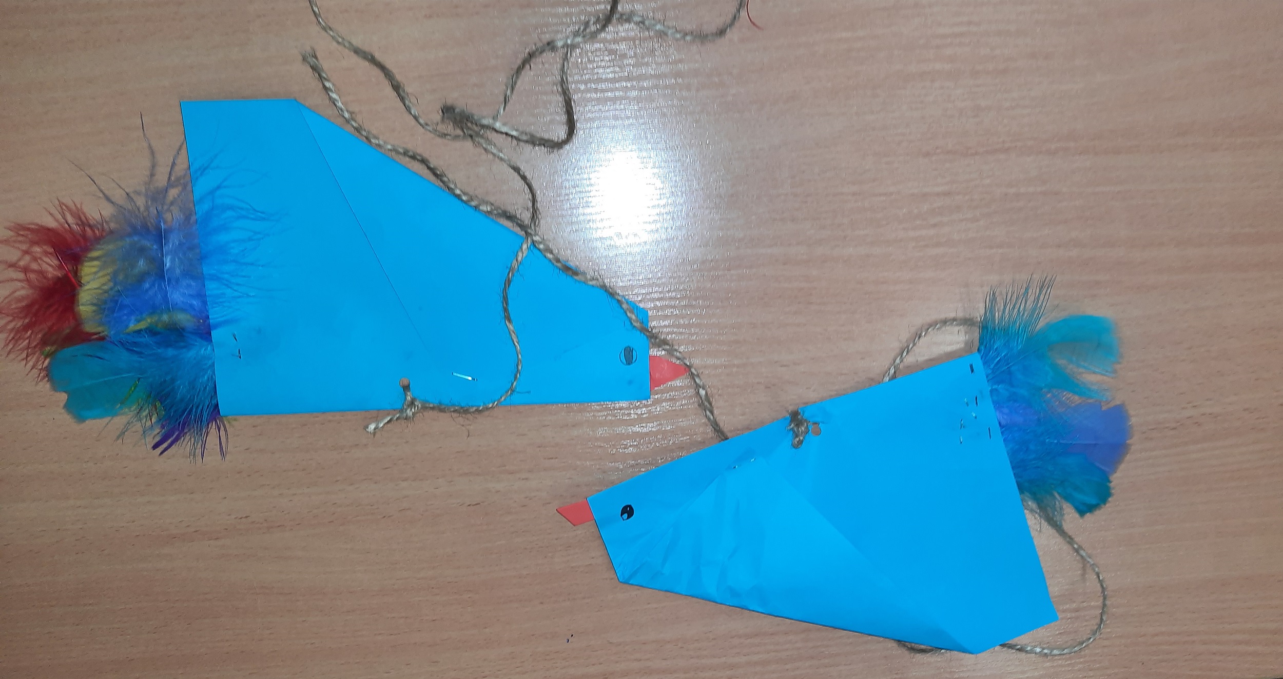 Na stoliku leżą dwa niebieskie latawce w kształcie ptaków z doklejonymi kolorowymi piórkami. Latawce również mają doczepione sznurki.