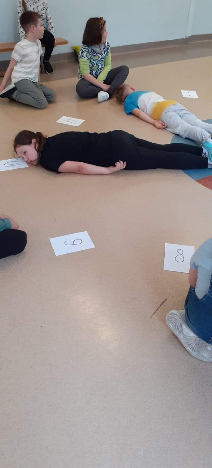 Na zdjęciu widać dzieci podczas zabawy "Żywy zegar". Na podłodze leży tarcza zegara ( kartki papieru z cyframi od 1 do 12). Dwoje dzieci leżą na podłodze jako wskazówki godziny.