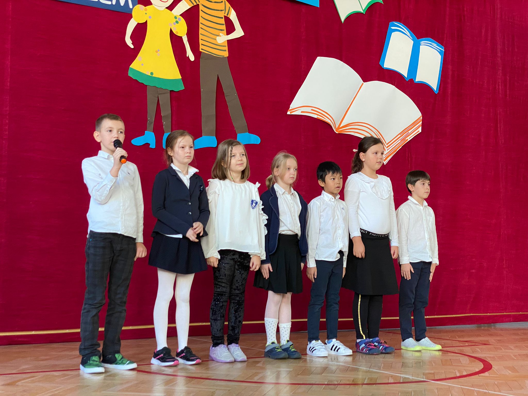 Dzieci ubrane na galowo stoją w szeregu. Jedno z dzieci ma mikrofon. Za nimi wisi kotara z przyczepioną ogromną książką.