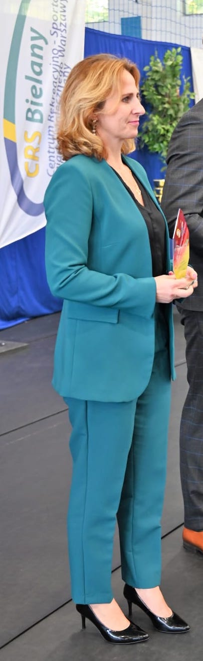 Krawiec, ubrana w turkusowy garnitur i buty na szpilkach, trzyma statuetkę Szkolnego Koła Zawodów Sportowych.