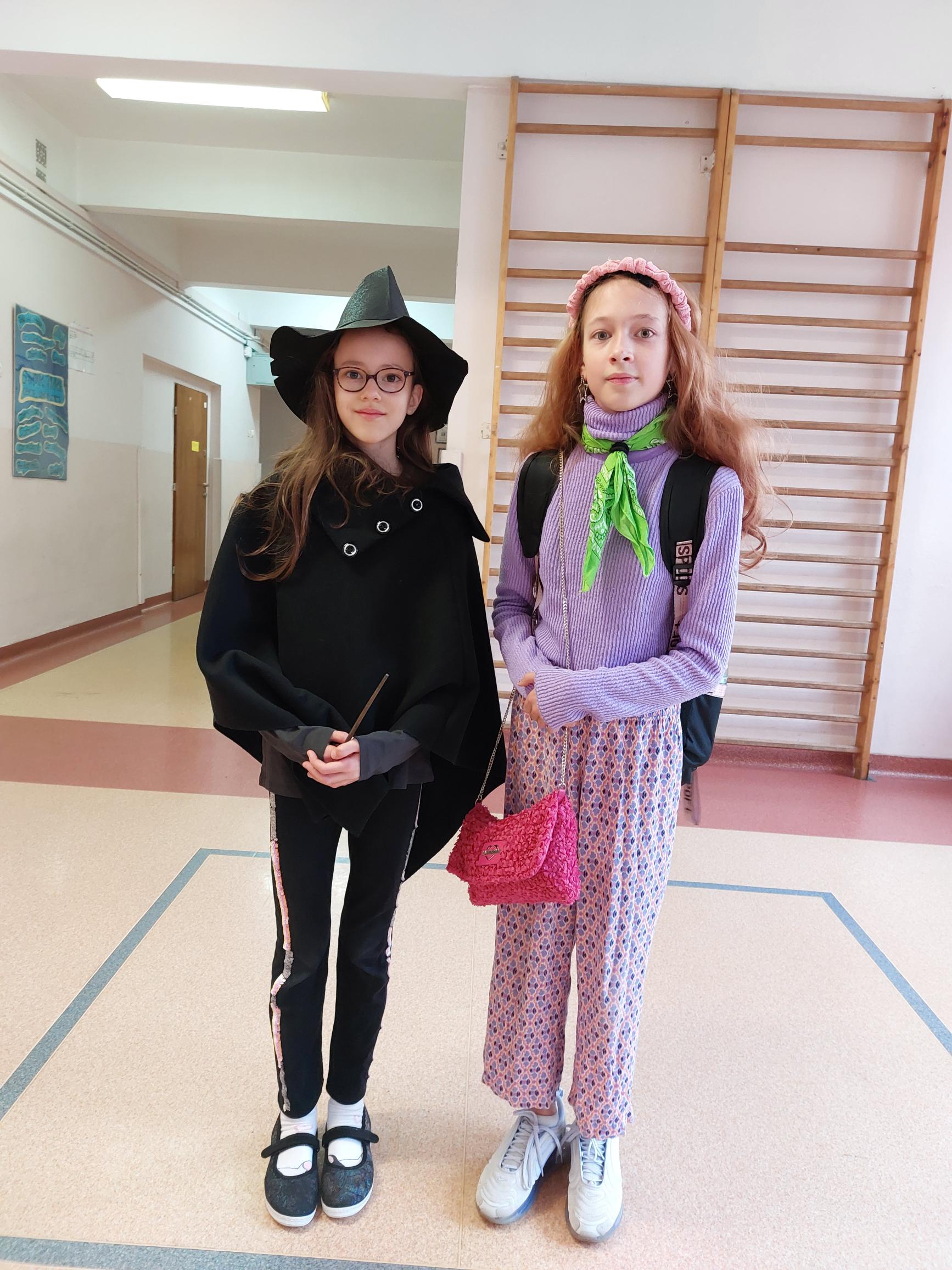 Dwie dziewczynki stoją obok siebie. Jedna ma na sobie czarny kapelusz z szerokim rondem i długim szpicem. Druga trzyma w ręku różową torebkę.