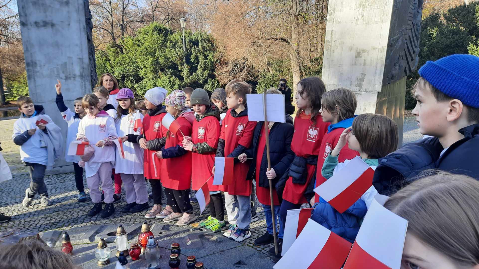 Dziewczynki i chłopcy w biało-czerwonych koszulkach z flagami Polski zapalają znicze na Pomniku Sapera.