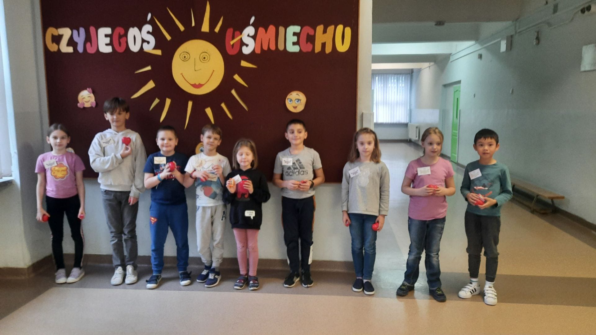 Cztery dziewczynki i pięciu chłopców z klas 1-3 wyróżnieni tytułem "Najżyczliwsi 2022" stoją przed tablicą. W rękach trzymają czerwone serduszka.