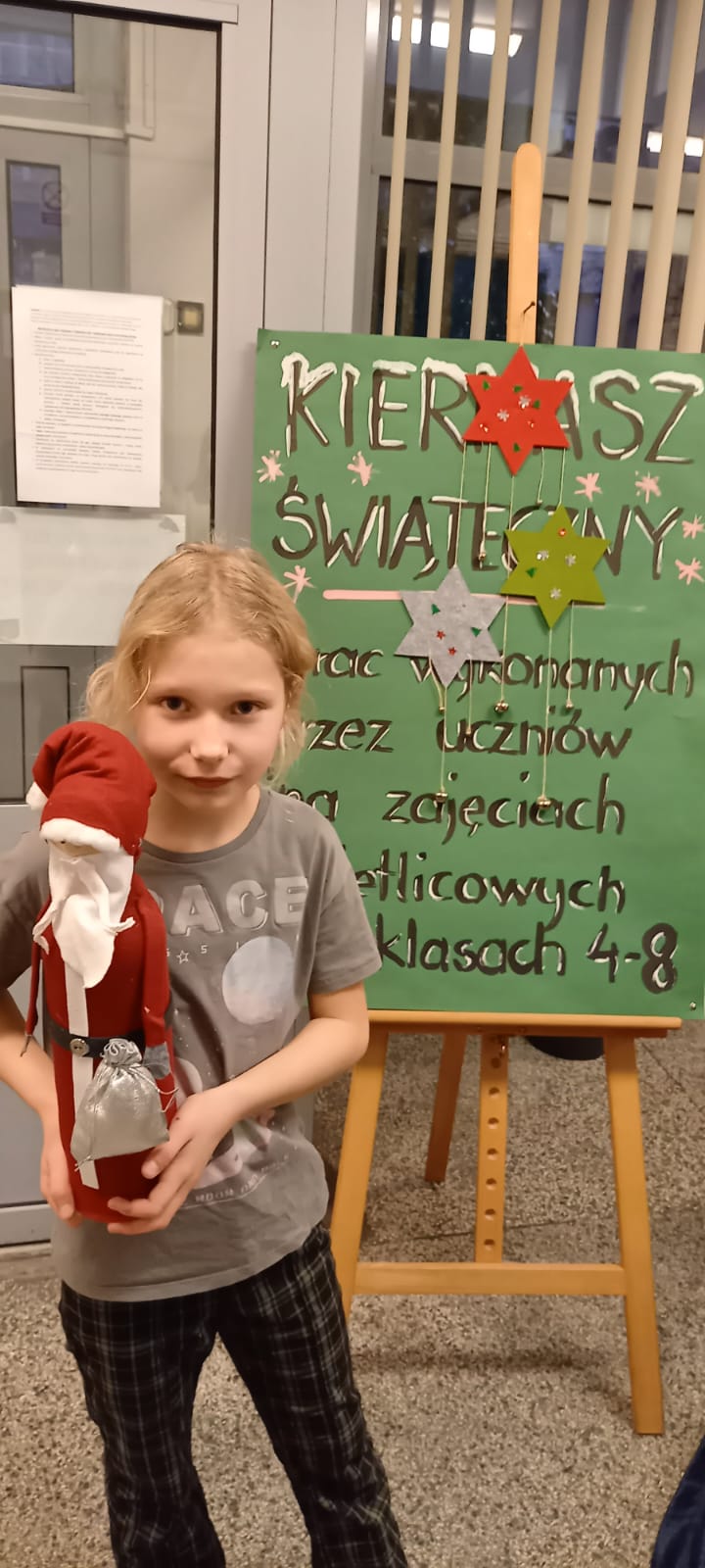 Uczennica z błąd włosami trzyma w ręku Mikołaja. Za nią stoi tablica informująca o kiermaszu bożonarodzeniowym.