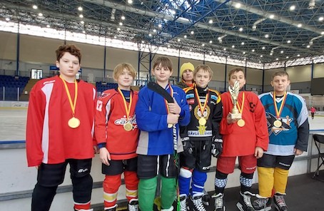 Sześciu chłopców wraz z trenerką stoi przed lodowiskiem. Mają na sobie stroje hokejowe a na piersiach złote medale. Jeden z zawodników trzyma statuetkę.