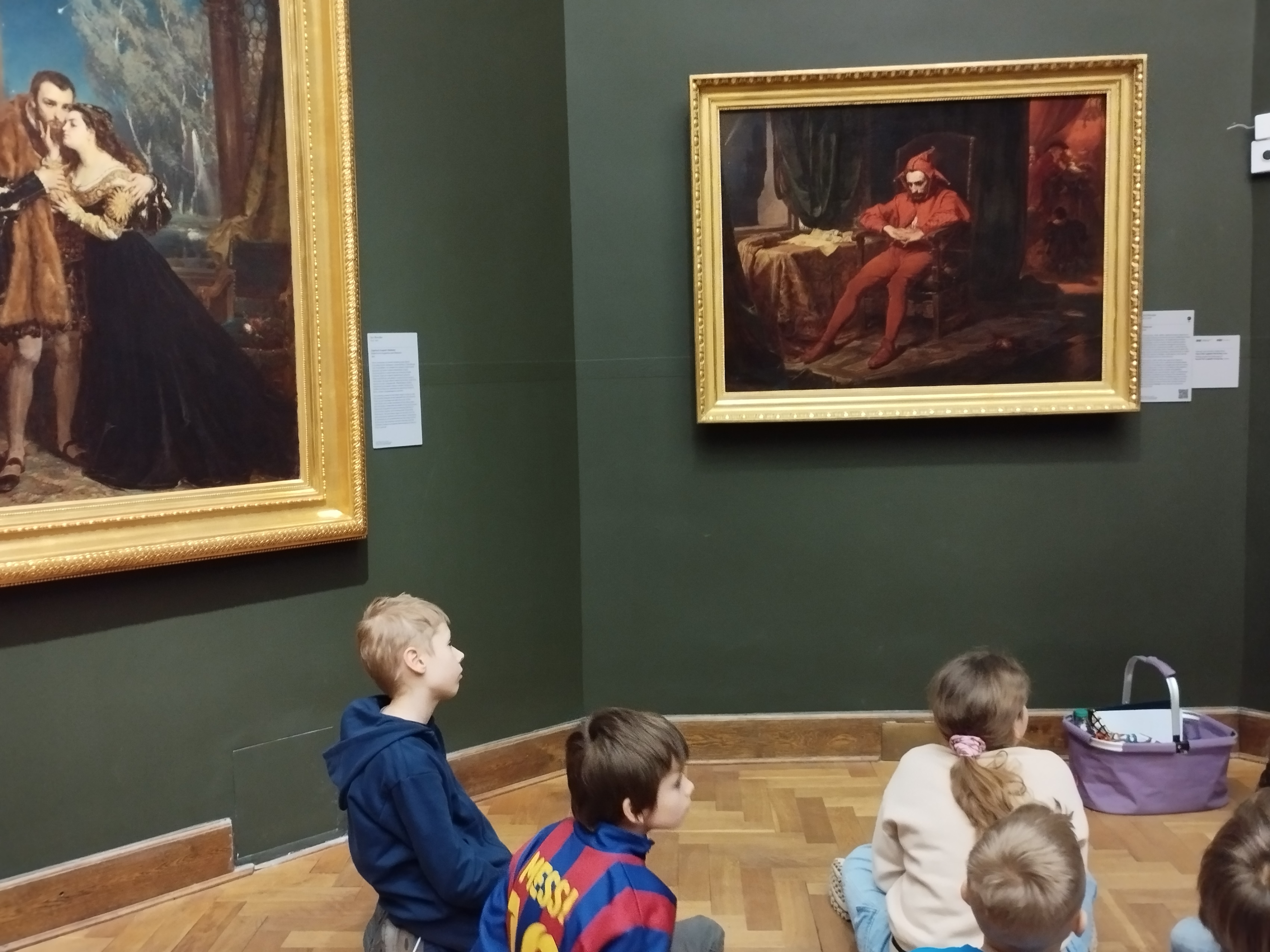 Ucznioowie w sali muzealnej siedzą na podłodze. Widzimy ich sylwetki z profilu lub wzrócone tyłem. Przed nimi na ścianach wisza obrazy w złotych ramach.