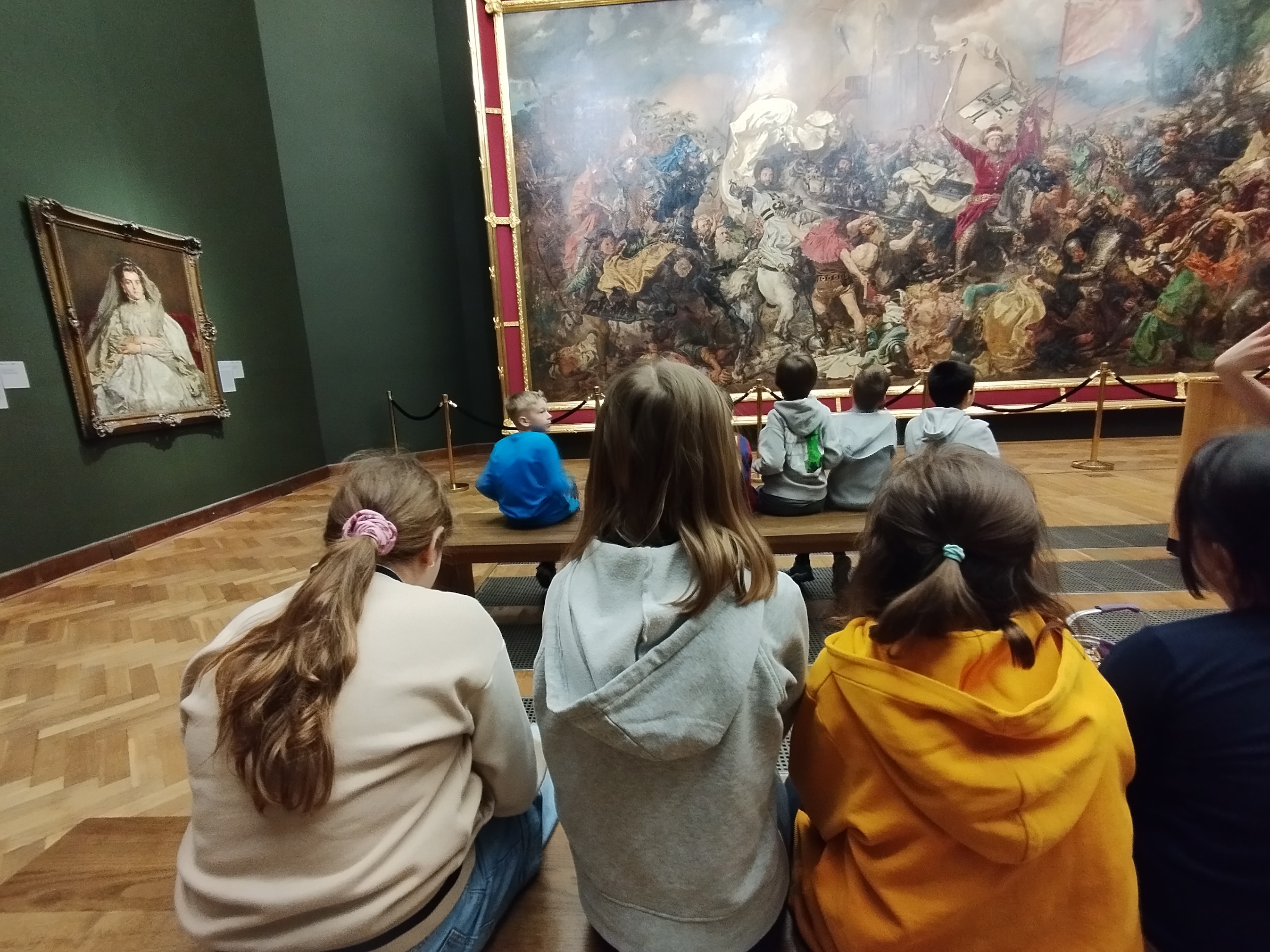W sali muzealnej uczniowie siedzą na ławkach. Są zwrócemi plecami. W głębi przed nimi wisi duży obraz w złotej ramie.