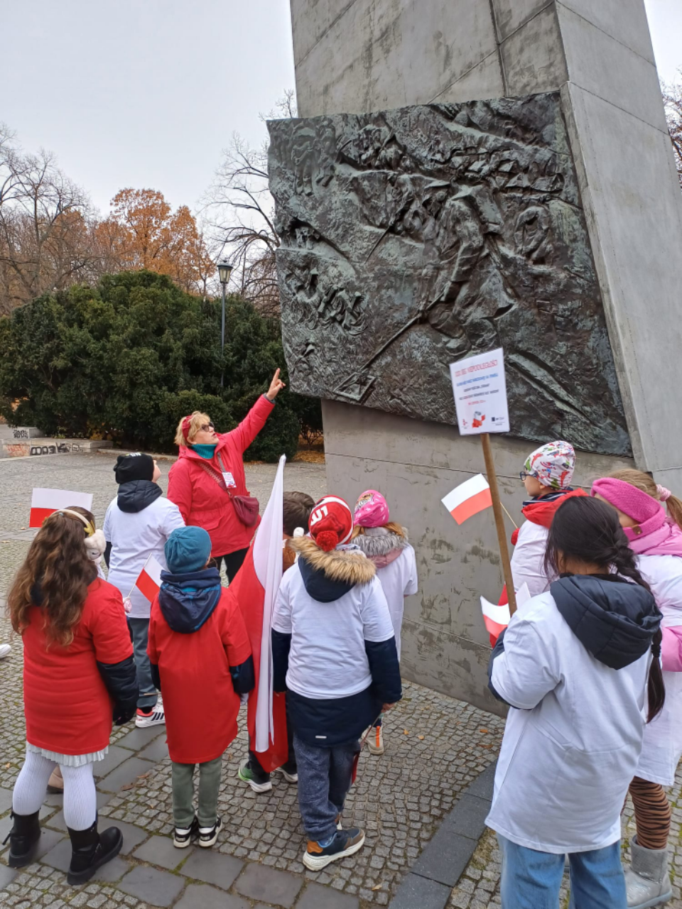 Pani w czerwonej kurtce opowiada dzieciom trzymającym flagi w białych i czerwonych koszulkach o historii pomnika Sapera.