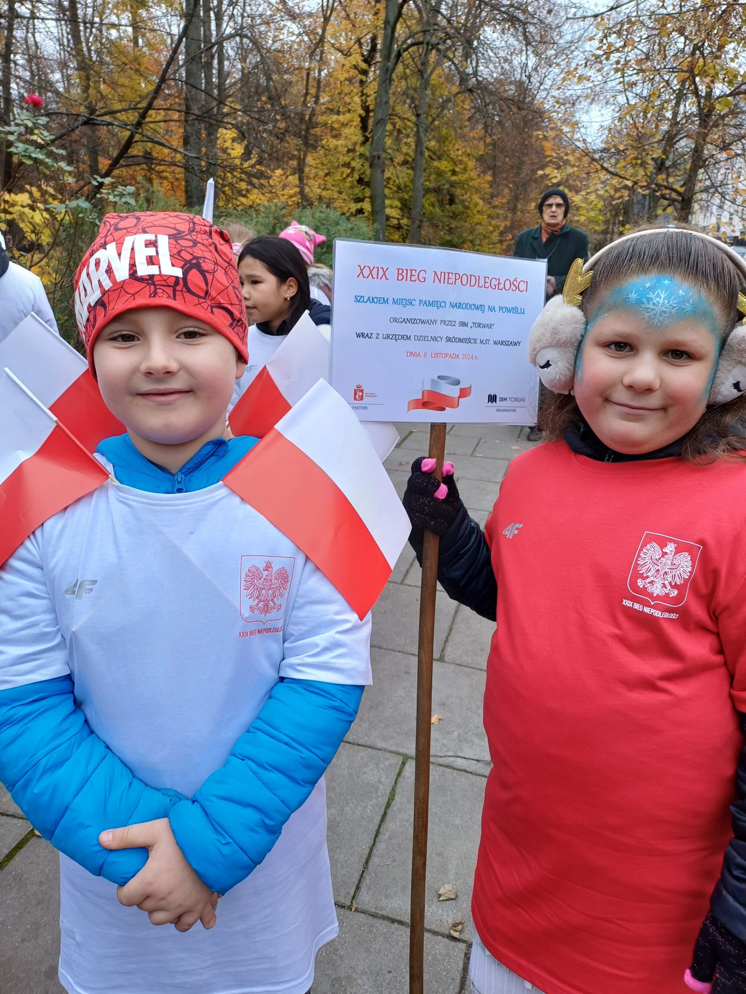 Chłopiec i dziewczynka z klasy 2b. Dziewczynka w ręku trzyma tabliczkę z informacją o Biegu. Chłopiec ma biało czerwone flagi, czerwoną czapkę i białą bluzkę.