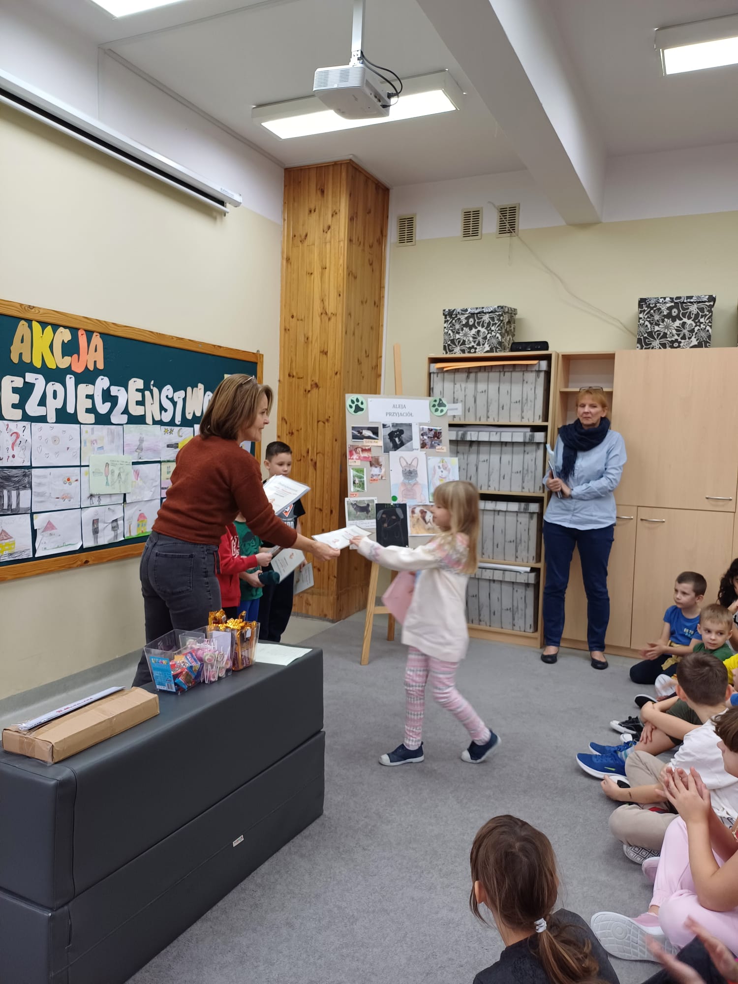 Dziewczynka podchodzi do nauczycielki po odbiór podziękowania za udział w konkursie Portret kundelka. Z tyłu tablica z rysunkami wykonanymi przez dzieci. Z przodu dzieci biją brawo.