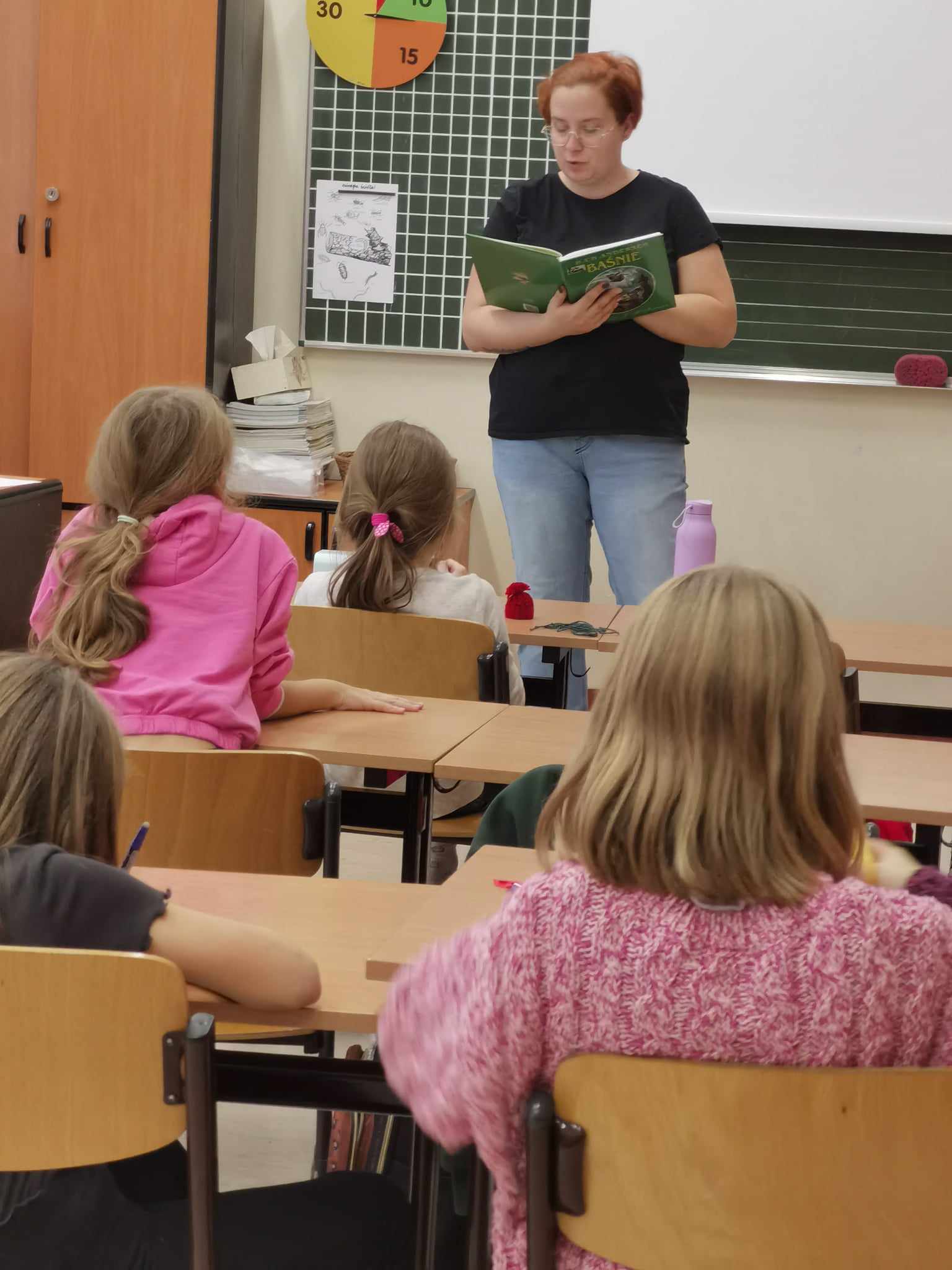 Uczniowie siedzą przy stolikach, przed nimi stoi kobieta ubrana w czarną koszulkę w rękach trzyma książkę.