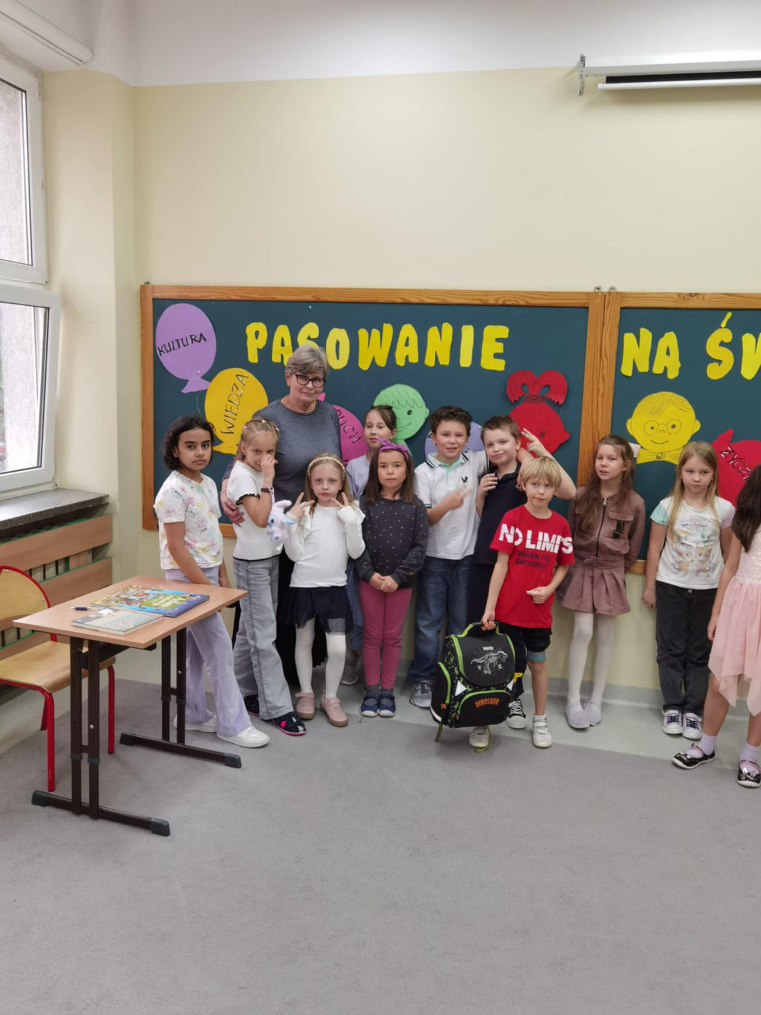 Na tle gazetki ściennej z żółtym napisem Pasowanie oraz kolorowymi balonikami stoi pani dyrektor w otoczeniu uśmiechniętych dzieci. Z lewej strony zdjęcia widoczny stolik z otwartą książką i wsuniętym krzesłem.