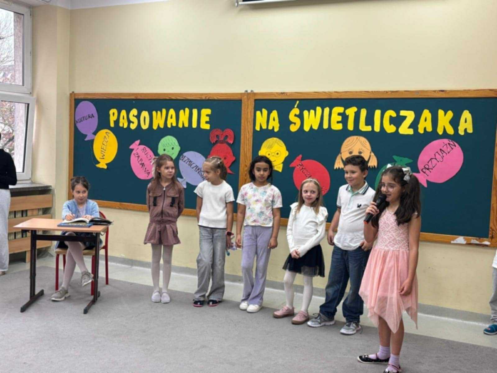 Na tle napisu 'Pasowanie na świetliczaka' z lewej strony zdjęcia widać dziewczynkę siedzącą przy stoliku i książkę. Obok niej 6 dzieci.