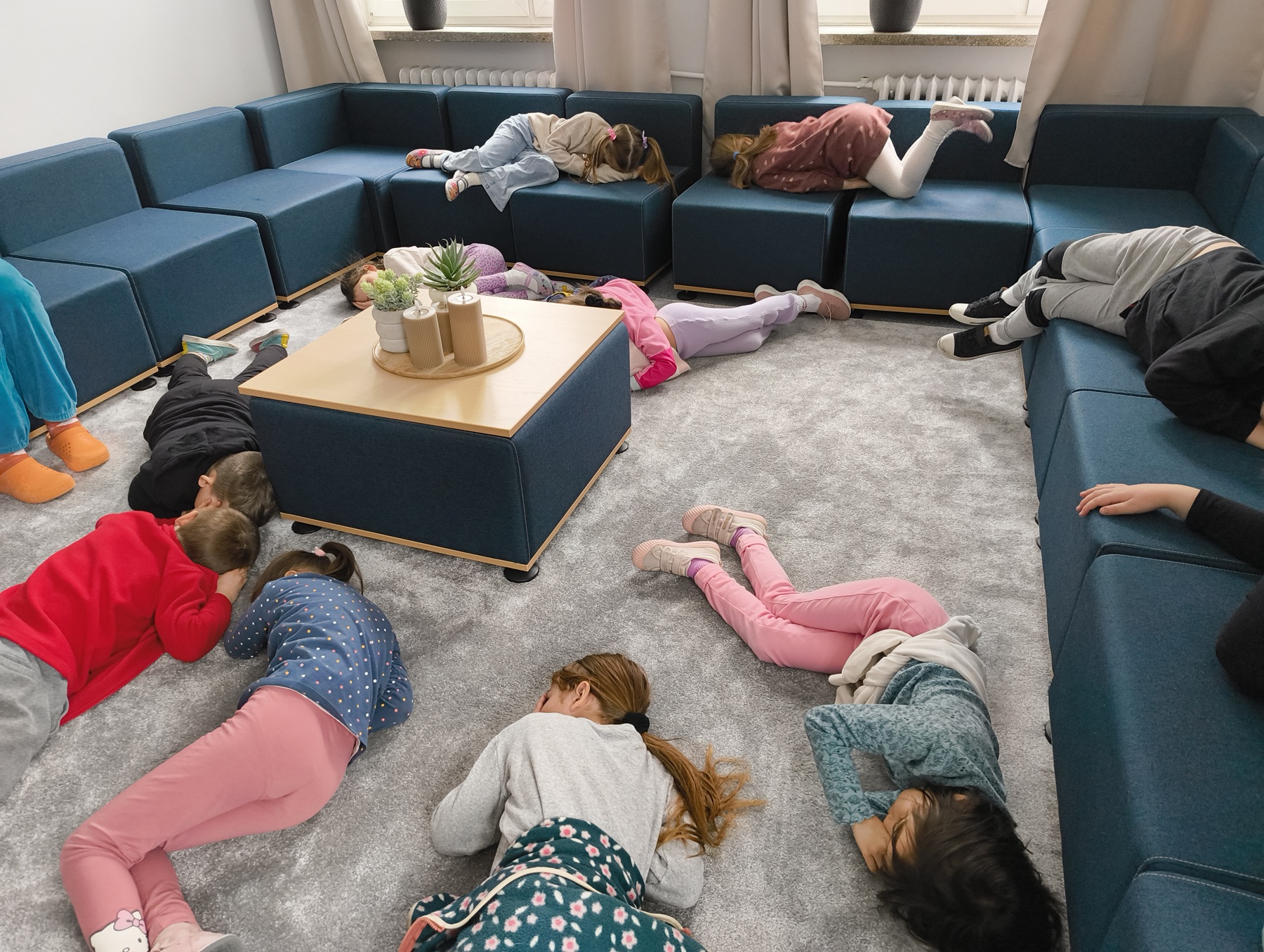 2.Na zdjęciu widać grupę dzieci przebywających w przytulnej, nowoczesnej sali relaksu. Pomieszczenie wyposażone jest w miękkie, modułowe kanapy ustawione wzdłuż ścian oraz niski stolik z dekoracyjnymi roślinami pośrodku. Na podłodze znajduje się miękki dywan, kt&oacute;ry sprzyja odpoczynkowi i wyciszeniu.
