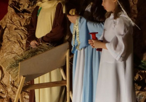 W stajence betlejemskiej stoją Maryja, Józef i Anioł. Maryja w swoich ramionach trzyma figurkę Dzieciątka Jezus.