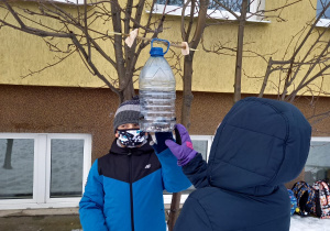 Dwoje dzieci zawiesza na gałęzi karmnik z plastikowej butelki.