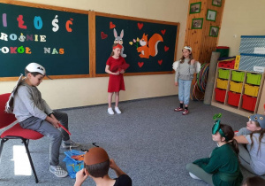 Na zdjęciu widać scenkę: chłopczyk gra rolę żurawia ( siedzi na krześle i łowi ryby) dziewczynka -rolę czapli w inscenizacji wiersza Jana Brzechwy „Żuraw i czapla” Na środku stoi narrator.