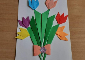 Fotografia przedstawia bukiet kwiatów wykonanych techników origami. Kwiaty połączone kokardką i przyklejone na białej kartce. Autorka: Tosia Mirosław kl.3b.