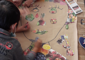 Na podłodze leży duży arkusz papieru z narysowanym konturem twarzy. Uczniowie przyklejają wycięte z gazetek zdjęcia owoców i warzyw.