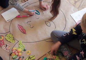 Na podłodze leży duży arkusz papieru z narysowanym konturem twarzy. Uczniowie dorysowują i kolorują oczy.