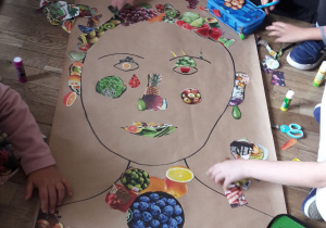 Na podłodze leży duży arkusz papieru z narysowanym konturem twarzy. Uczniowie przyklejają wycięte z gazetek zdjęcia owoców i warzyw.