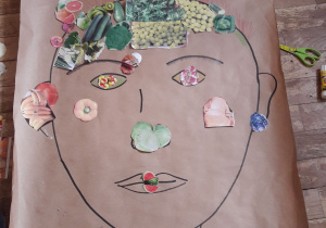 Na podłodze leży duży arkusz papieru z narysowanym konturem twarzy. Uczeń rysuje ozdobę na szyi.
