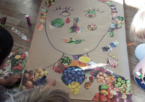 Na podłodze leży duży arkusz papieru z portretem wyklejonym zdjęciami owoców i warzyw