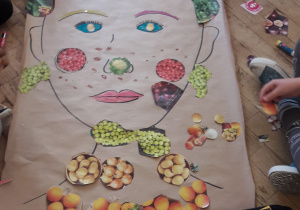 Na podłodze leży duży arkusz papieru z portretem wyklejonym zdjęciami owoców i warzyw.