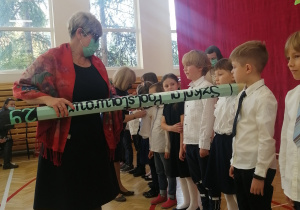 Pierwszaki stoją w rzędzie twarzą do widowni. Pani Dyrektor pasuje dzieci na uczniów dotykając ramienia wielkim, zielonym ołówkiem.