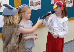 Dziewczynka z klasy 1b przebrana za koguta recytuje tekst przez mikrofon. Obok niej stoją dwie dziewczynki przebrane za myszki.