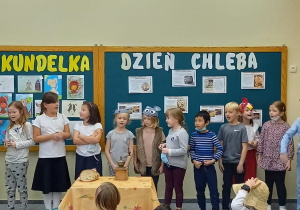 Dzieci z klas 1-3 oglądają inscenizację Kogut i dwie myszki w wykonaniu uczniów z klasy 1b. Na środku stoi stolik przykryty obrusem na którym leży chleb. W tle dekoracja z okazji Dnia chleba.