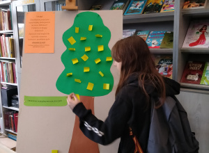 Uczennica przykleja na drzewie karteczkę z propozycją zakupu książki do biblioteki.