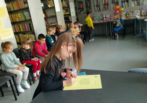 Dziewczynka przy szarym stoliku podpisuje tekst przysięgi. Za nią siedzą w półkole pozostali uczniowie z klasy 1a.
