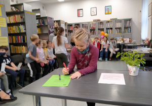 Dziewczynka przy szarym stoliku podpisuje tekst przysięgi. Za nią siedzą w półkole pozostali uczniowie z klasy 1b.