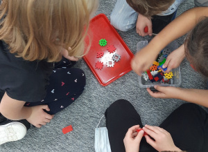 Kilkoro uczniów siedzi na dywanie. Przed nimi położona jest czerwona taca i bezbarwne pudełko plastikowe, a w nich kolorowe klocki.