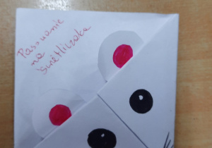 Na zdjęciu jest zakładka do książki wykonana techniką origami. Przedstawia ona myszkę. Zrobiona jest z białego papieru i ma kształt kwadratu. Pyszczek i oczy są namalowane czarnym markerem. Uszy są białe, okrągłe a środku namalowana różowa kropka.