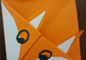Zdjęcie przedstawia zakładkę do książki wykonaną techniką origami. Zakładka przedstawia liska. Ma formę kwadratu. Jest pomarańczowa z białymi elementami na pyszczku i uszach. Oczy i nosek narysowane markerem.