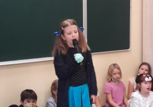 Dziewczynka śpiewa piosenkę. Ubrana jest w niebieską spódniczkę i czarna bluzkę z białymi koreczkami. Włosy ma uczesane w dwa kucyki z niebieskimi kokardami.