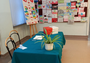 Na przykrytym zielonym suknem stole stoi biało-czerwona urna. Obok znajduje się kwiatek doniczkowy. W tle widoczna jest tablica z pracami plastycznymi.