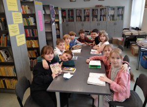 Pięć dziewczynek i 6 chłopców siedzi wokół dużego stołu w bibliotece szkolnej. Przed każdym z uczniów na stole leży otwarta książka. Dzieci patrzą w obiektyw.