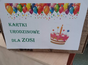 Na stoliku stoi pudełko z napisem na białym tle "Kartki urodzinowe dla Zosi. Nad napisem zawieszone są kolorowe balony. Po prawej stronie napisu jest różowy tort.