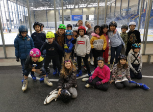 Klasa 3a. Uczniowie w kasach i łyżwach stoją przed bandą na lodowisku.