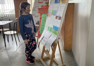 Przy ścianie korytarza stoją sztalugi z wystawą prac uczniowskich na temat frazeologizmów. Przed nimi dziewczynka z kucykiem, w maseczce na nosie i ustach ubrana w kolorowe spodnie oraz bluzkę z jednorożcem przygląda się rysunkom.