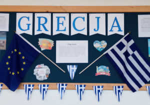 Na granatowej tablicy zamieszczony jest napis Grecja. Obok znajduje się flaga Grecji i Unii Europejskiej.