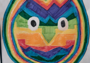 Praca plastyczna przedstawia pisankę pomalowaną flamastrami. Kolory użyte tu to: niebieski, zielony, pomarańczowy,żółty, czerwony. Pisanka ozdobiona jest na kształt ludzkiej twarzy. Ma czarne oczy, widoczny nos i usta.