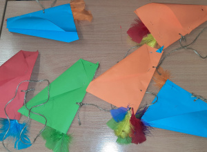 Na stoliku leżą kolorowe latawce w kształcie ptaków (czerwone,niebieskie, zielone, pomarańczowe). Latawce wykonane są z papieru i mają doklejone kolorowe sztuczne piórka.
