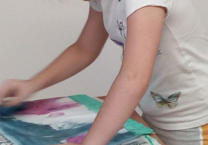 Zdjęcie przedstawia pochyloną dziewczynkę, brunetkę z końskim ogonem. Jest ona oparta na ławce pokrytej gazetami, stoją na niej farby plakatowe w pojedynczych słoiczkach, przed nią leży praca, którą z zapałem maluje.
