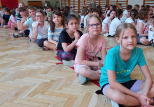 Uczniowie siedzą na podłodze w rzędach w sali gimnastycznej.