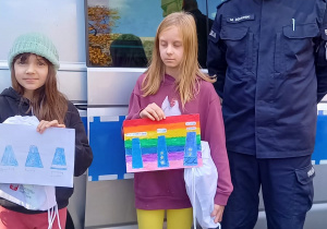 Pan Policjant i dwie uczennice stoją przed radiowozem. Dziewczynki w rękach trzymają prace plastyczne przedstawiające znaczki odblaskowe.